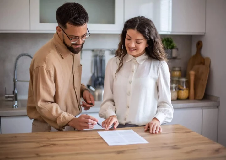 Imagem de um homem e uma mulher em uma cozinha, sorrindo e analisando um documento juntos em um balcão de madeira. O homem está segurando uma caneta, pronto para assinar, enquanto a mulher aponta para o papel