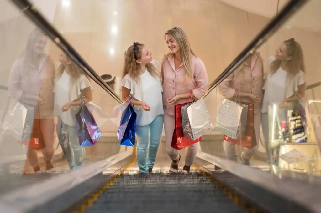 Imagem de duas mulheres na escada rolante de um shopping center com sacolas de compras nos braços para ilustrar matéria sobre o que fazer no Rio com chuva