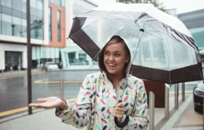 Imagem de uma mulher com casaco e um guarda chuva andando na rua de uma cidade enquanto chove para ilustrar matéria sobre o que fazer no Rio de Janeiro com chuva