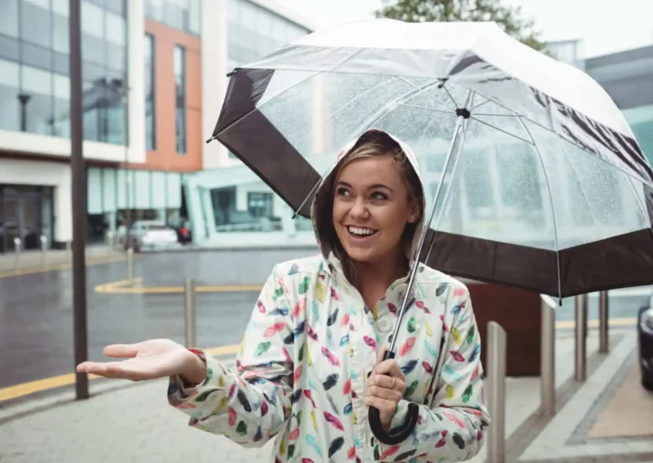 Imagem de uma mulher com casaco e um guarda chuva andando na rua de uma cidade enquanto chove para ilustrar matéria sobre o que fazer no Rio de Janeiro com chuva