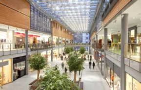 Imagem de shopping moderno e espaçoso com dois andares, teto de vidro, árvores internas e lojas em ambos os andares, com luz natural preenchendo a área central aberta enquanto as pessoas caminham e fazem compras para ilustrar matéria sobre os melhores shoppings de SP