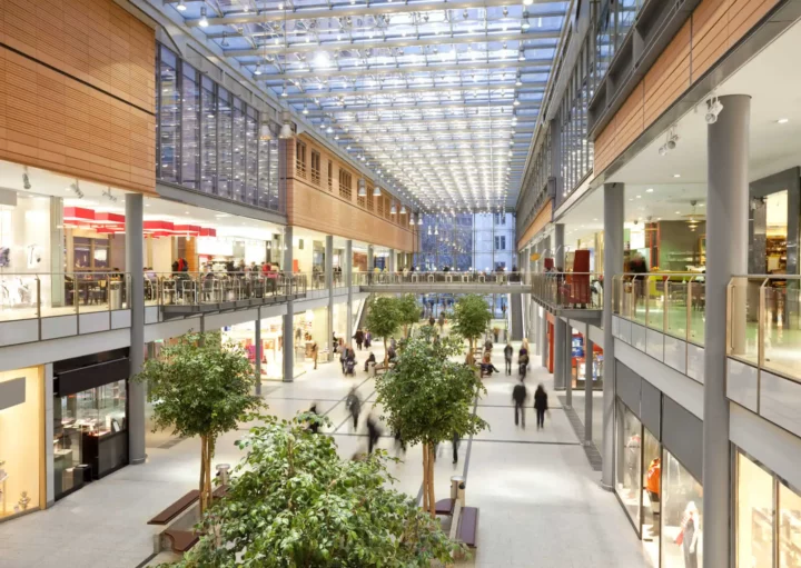 Imagem de shopping moderno e espaçoso com dois andares, teto de vidro, árvores internas e lojas em ambos os andares, com luz natural preenchendo a área central aberta enquanto as pessoas caminham e fazem compras para ilustrar matéria sobre os melhores shoppings de SP