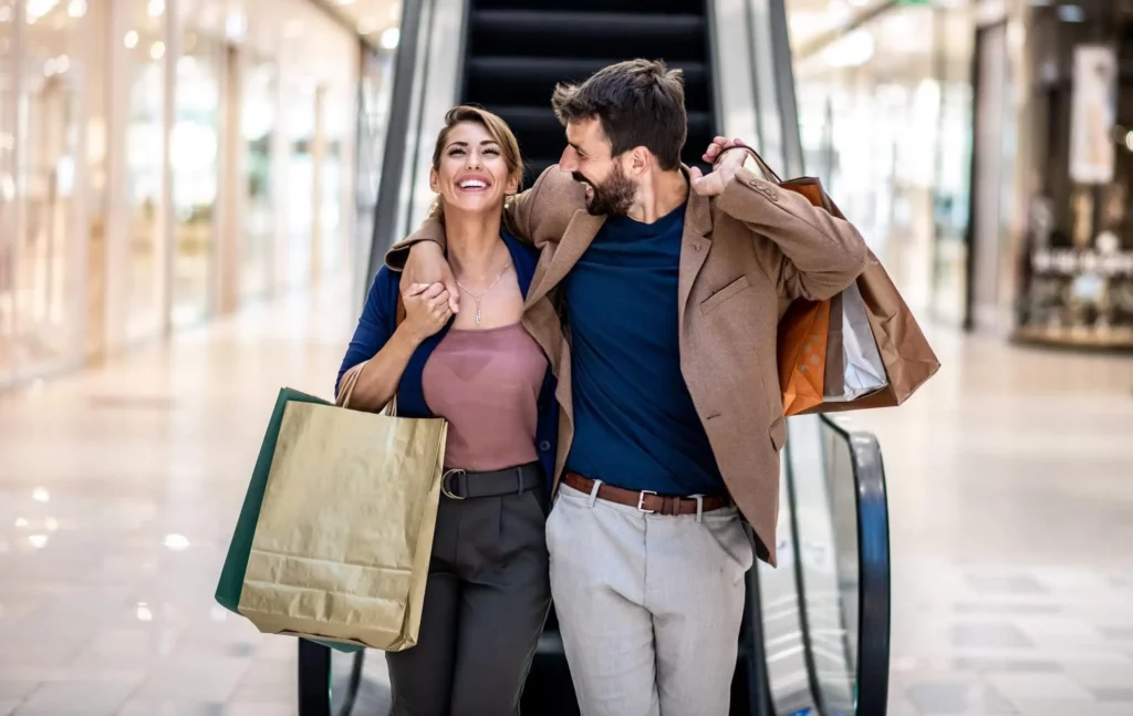 Imagem de um casal sorridente segurando sacolas de compras caminha de braços dados em um shopping center, parado em frente a uma escada rolante. Eles parecem felizes e bem vestidos para ilustrar matéria sobre qual o shopping de luxo de São Paulo