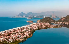 Foto que ilustra matéria sobre a Região Oceânica de Niterói mostra uma vista do alto dos bairros de Piratininga e Itaipu, com a Lagoa de Piratininga à direita, casas e prédios ao centro, a praia à esquerda e as cidades de Niterói e do Rio de Janeiro ao fundo. (Crédito: Getty Images)