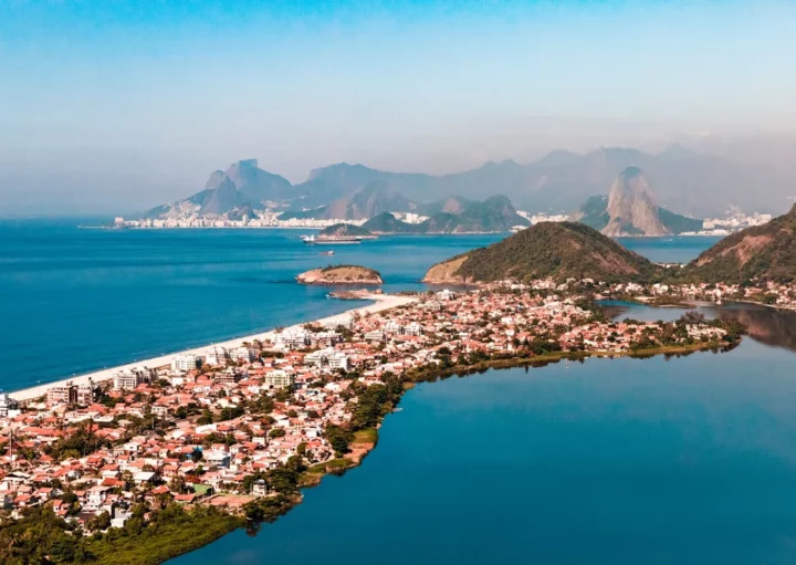 Foto que ilustra matéria sobre a Região Oceânica de Niterói mostra uma vista do alto dos bairros de Piratininga e Itaipu, com a Lagoa de Piratininga à direita, casas e prédios ao centro, a praia à esquerda e as cidades de Niterói e do Rio de Janeiro ao fundo. (Crédito: Getty Images)