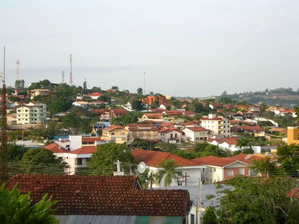 Imagem de uma vista panorâmica em Águas de São Pedro, em São Paulo, mostra casas com telhados vermelhos, cercada por árvores e vegetação,prédios mais altos e torres de comunicação sob um céu claro