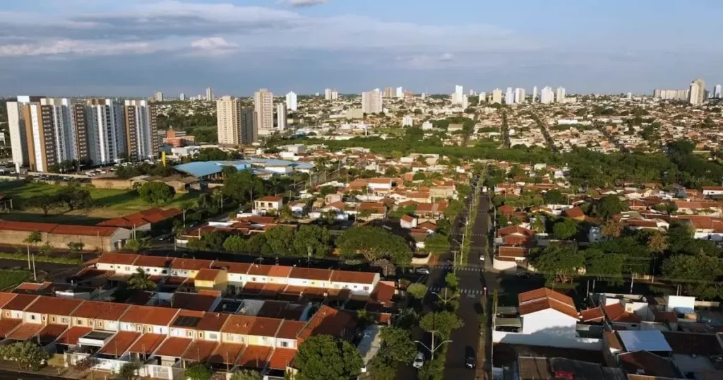 Imagem da vista aérea de Araraquara mostra casas baixas com telhados vermelhos e prédios de apartamentos altos, árvores verdes e estradas que se estendem à distância sob um céu parcialmente nublado
