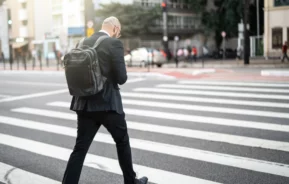 Imagem de um homem de costas vestindo terno preto com uma mochila nos ombros cruzando uma faixa de pedestres na Avenida Paulista para ilustrar matéria sobre o centro expandido de São Paulo