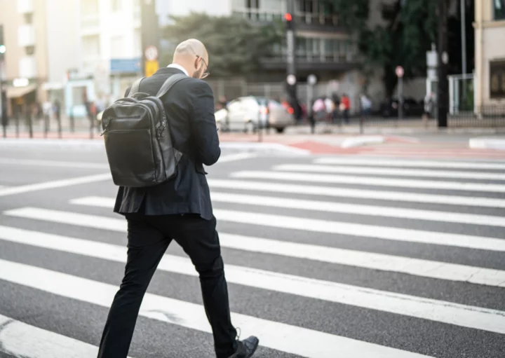 Imagem de um homem de costas vestindo terno preto com uma mochila nos ombros cruzando uma faixa de pedestres na Avenida Paulista para ilustrar matéria sobre o centro expandido de São Paulo