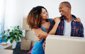 Imagem de um casal composto por um homem e uma mulher sorrindo um para o outro. O homem está segurando uma caixa de papelão para ilustrar matéria sobre como escolher uma cidade para morar