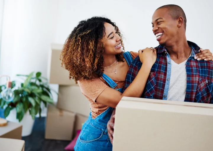 Imagem de um casal composto por um homem e uma mulher sorrindo um para o outro. O homem está segurando uma caixa de papelão para ilustrar matéria sobre como escolher uma cidade para morar