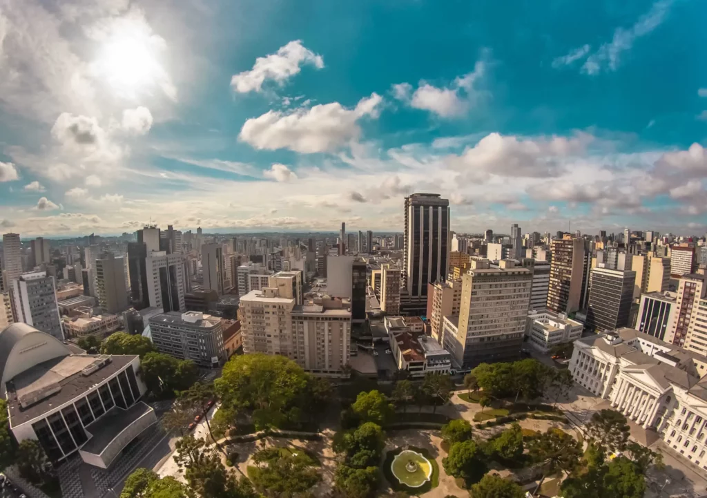 Imagem da vista aérea da praça Santos Andrade, no Centro de Curitiba, mostra vegetação e prédios ao redor em um dia de céu azul 