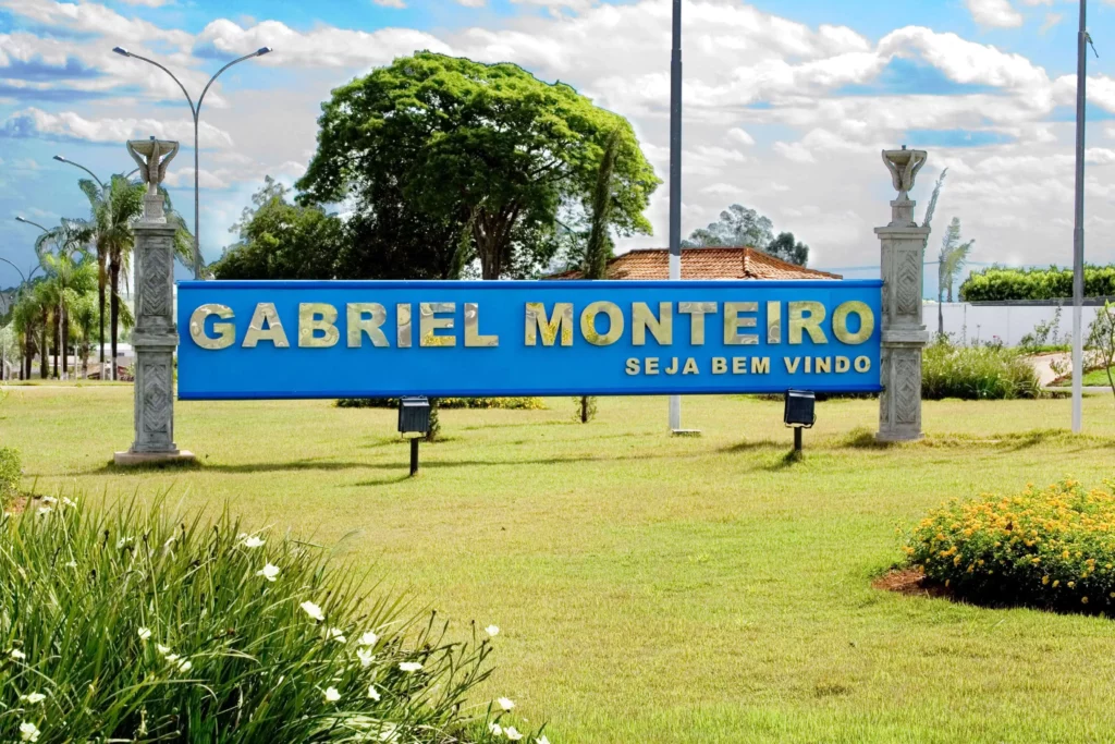 Imagem de uma grande placa azul em uma área gramada diz Gabriel Monteiro Seja Bem-Vindo, dando as boas-vindas aos visitantes com árvores, flores e uma casa ao fundo sob um céu parcialmente nublado