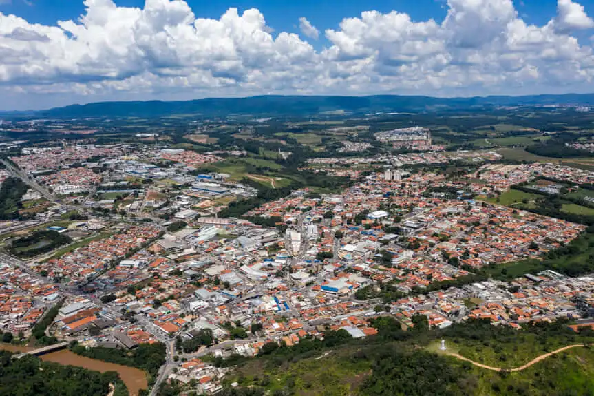 Imagem da vista aérea de Itupeva, em SP, mostra casas, prédios e ruas densamente povoadas, cercadas por campos verdes e colinas sob um céu parcialmente nublado
