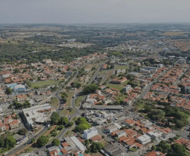 Imagem da vista aérea de Jaguariúna, em SP, mostra casas, espaços verdes e estradas que serpenteiam pela vizinhança, cercada por árvores e terrenos abertos sob um céu nublado