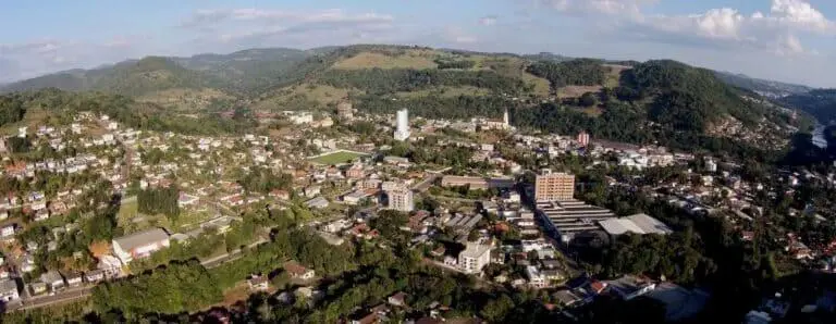 Imagem da vista aérea de Luzerna mostra a cidade cercada por colinas verdes e florestas, com aglomerados de edifícios, estradas e espaços abertos sob um céu parcialmente nublado