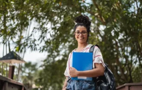 Estudante na frente de uma árvore, segurando apostilas e com uma bolsa nas costas.