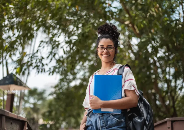 Estudante na frente de uma árvore, segurando apostilas e com uma bolsa nas costas.