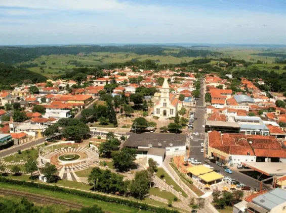 Imagem da vista aérea de Pompéia, em SP, mostra prédios de telhados vermelhos, uma igreja central, ruas arborizadas e uma praça circular cercada por campos verdes e colinas ondulantes sob um céu azul