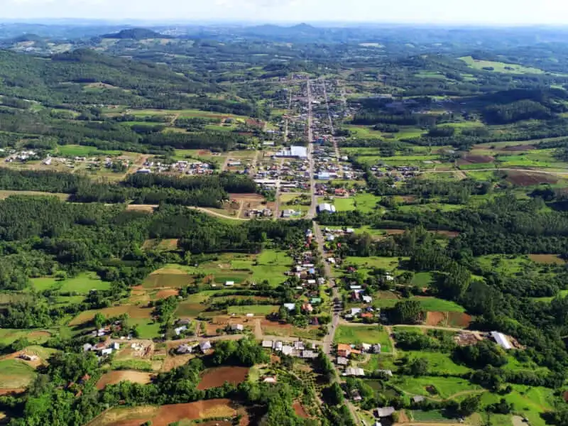 Imagem da vista aérea de Presidente Lucena, no RS, mostra casas espalhadas, terras agrícolas e campos verdes que se estendem à distância. Colinas arborizadas e céu azul são visíveis ao fundo