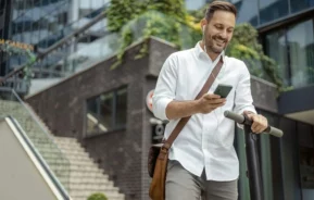 Imagem de um homem vestindo camisa branca e bolsa marrom a tiracolo sorri enquanto olha seu telefone e ouve fones de ouvido, andando em uma scooter elétrica em uma das cidades do Brasil com melhor qualidade de vida, cercada por edifícios modernos e escadas