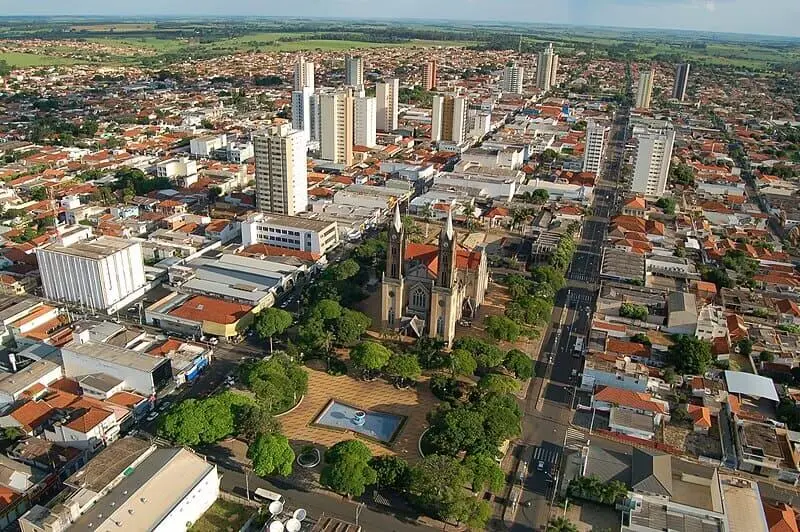Imagem da vista aérea de Votuporanga, em São Paulo, mostra prédios altos, casas baixas e uma praça central com uma igreja, árvores e uma fonte cercada por ruas movimentadas e infraestrutura urbana moderna