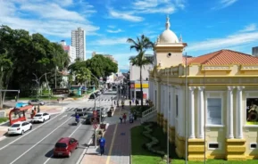Imagem de uma rua em São José dos Campos com carros, pessoas andando nas calçadas, palmeiras e um prédio histórico amarelo com uma cúpula. Prédios altos e um céu azul com nuvens esparsas completam a cena para ilustrar matéria sobre os bairros de São José dos Campos.