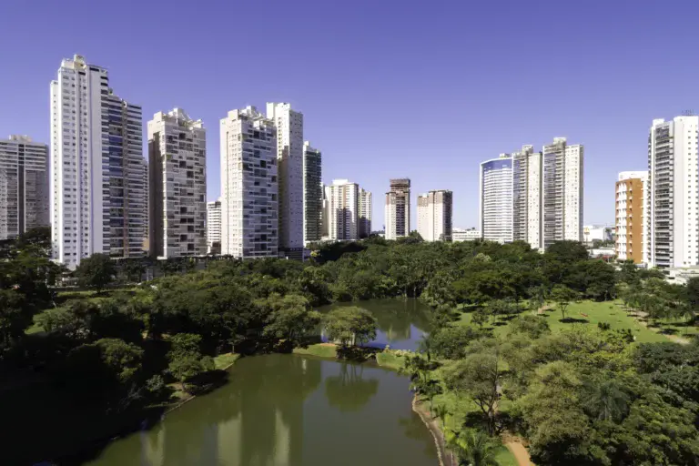 Imagem de prédios de apartamentos altos e modernos de Goiânia se alinham no horizonte atrás de um parque verde exuberante com árvores e um lago
