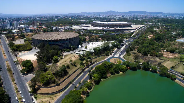 Imagem da vista aérea de dois grandes estádios esportivos em Belo Horizonte cercados por árvores e estradas, com um lago verde em primeiro plano e montanhas distantes ao fundo