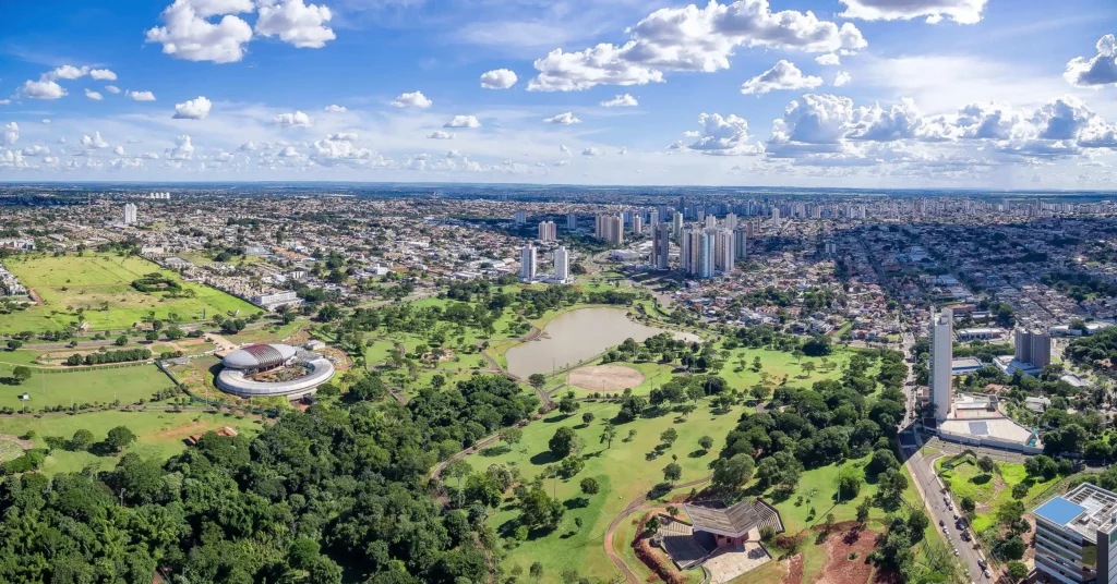  Imagem da aérea de Campo Grande (MS) mostra prédios altos, um parque central com muitas árvores, um lago e várias estruturas modernas sob um céu parcialmente nublado