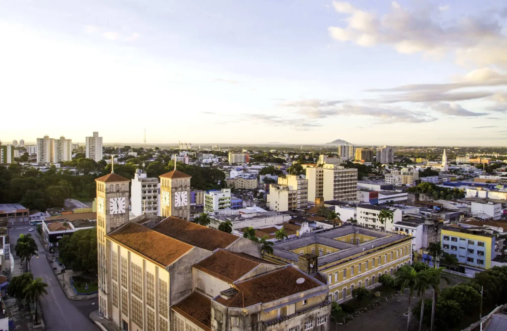 Imagem da vista aérea de Cuiabá ao pôr do sol mostra uma torre de relógio proeminente e um edifício histórico com telhado vermelho, cercado por edifícios modernos e vegetação exuberante