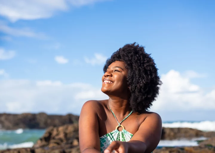 Imagem de uma mulher negra sorridente vestindo uma camisa azul sentada em uma praia olhando para o lado com um céu azul ao fundo para ilustrar matéria sobre as cidades mais seguras da Bahia