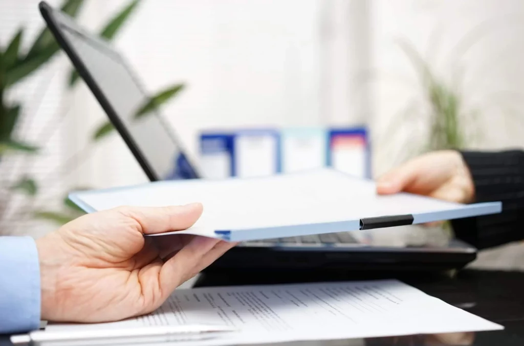 Duas pessoas trocam uma pasta sobre uma mesa com um laptop e documentos. Uma pessoa está entregando a pasta, enquanto a outra a recebe. O fundo está ligeiramente desfocado, com itens de escritório visíveis, para ilustrar matéria sobre como fazer um distrato de contrato de aluguel