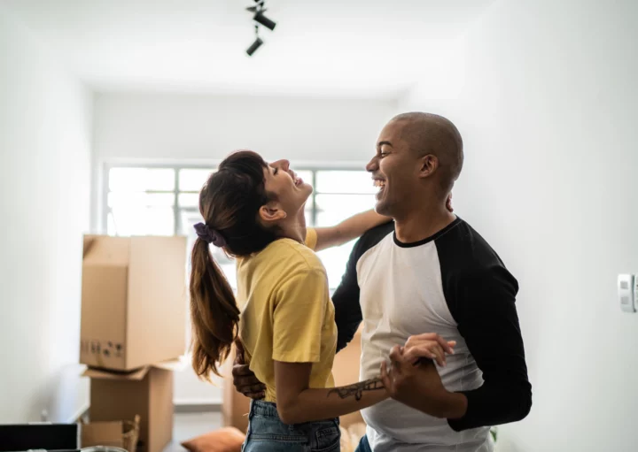 Imagem de um casal composto por um homem e uma mulher dançando com expressão de felicidade no rosto em sua casa nova para ilustrar matéria sobre como financiar 100% de um imóvel