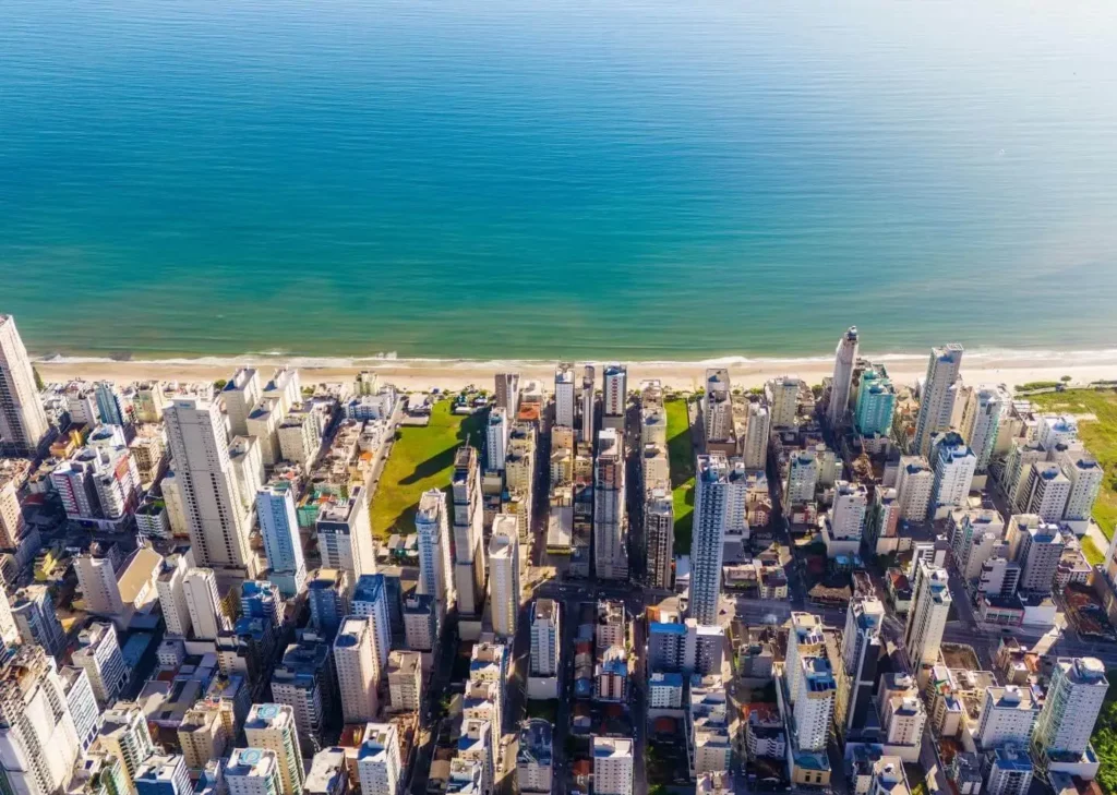 Imagem da vista aérea de uma cidade costeira em Santas Catarina densamente construída com prédios altos perto de uma praia de areia e um oceano azul calmo para ilustrar matéria sobre o custo de vida de Santa Catarina