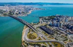 Imagem da vista aérea de uma cidade litorânea com pontes conectando áreas urbanas sobre águas azul-turquesa, cercada por edifícios, estradas e colinas verdes em Santa Catarina sob um céu azul claro.