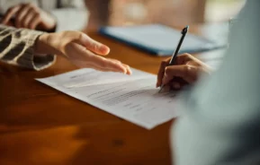 Close-up de duas pessoas em uma mesa; uma está segurando uma caneta e assinando um distrato de contrato, enquanto a outra gesticula em direção ao papel, explicando ou apontando algo no documento para ilustrar matéria sobre distrato de contrato de aluguel
