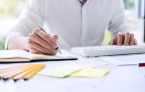 Imagem da silhueta de um homem com uma camiseta social branca sentado em uma mesa fazendo nota em um papel com uma caneta para ilustrar matéria sobre o financiamento de imóvel para MEI