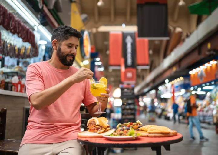 Imagem de um homem tomando uma bebida com alguns lanches em cima de uma mesa no Mercado Municipal para ilustrar matéria sobre os mercados em São Paulo