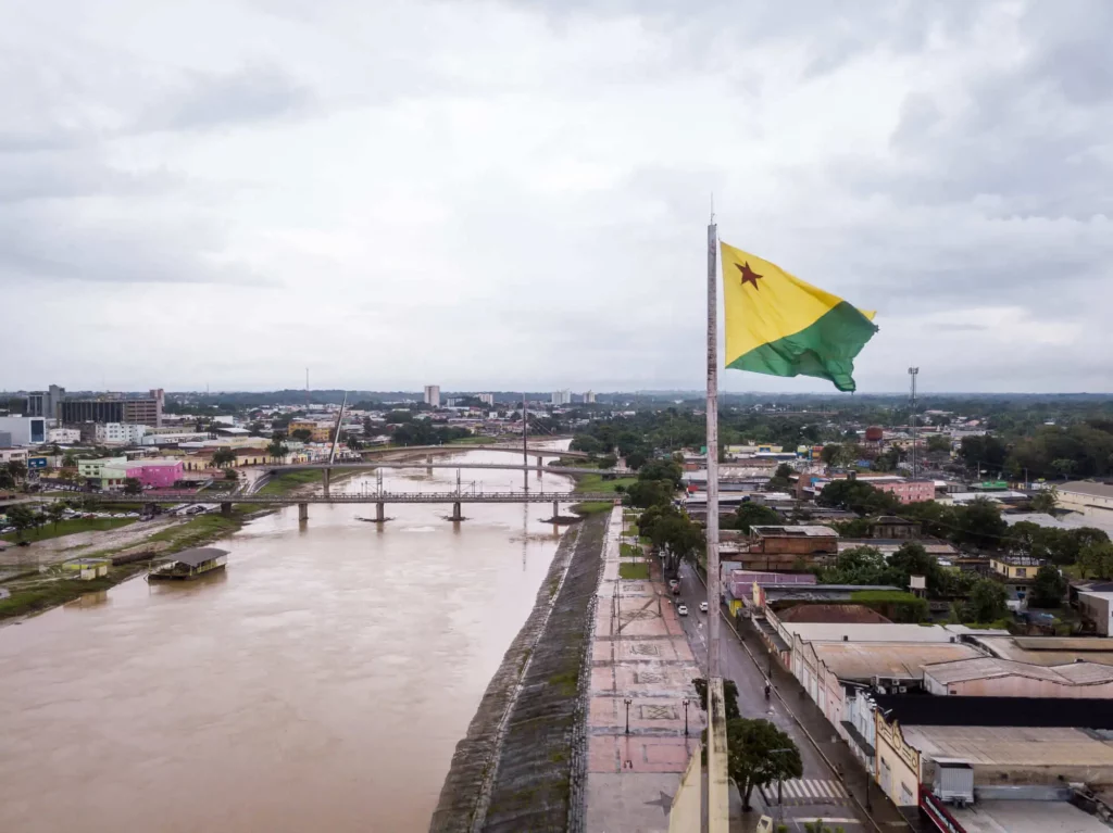 Imagem da bandeira do Acre estendida no alto da cidade de Rio Branco com vista para um rio