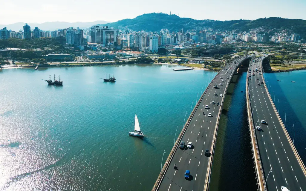 Imagem de vista aérea da cidade de Florianópolis, uma ponte com carros, veleiros na água e prédios com colinas ao fundo sob um céu claro para ilustrar matéria sobre a capital mais segura do Brasil