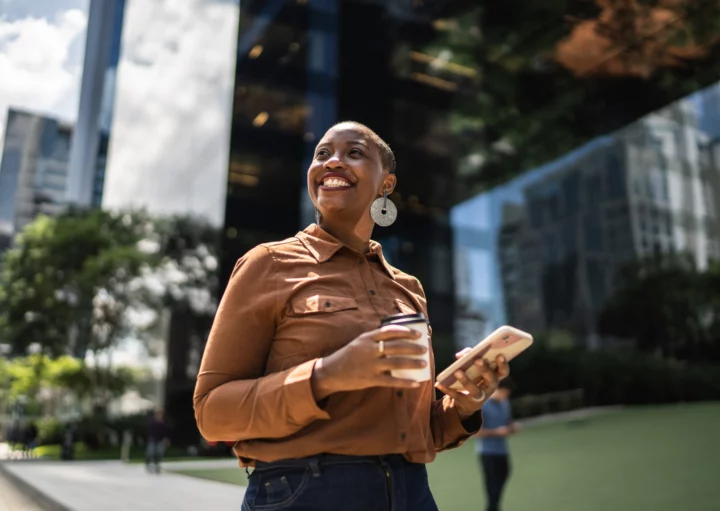 Imagem de uma mulher negra andando em uma rua em frente a um prédio empresarial, sorrindo e com um celular na mão, para ilustrar matéria sobre qual a cidade mais tranquila do Brasil