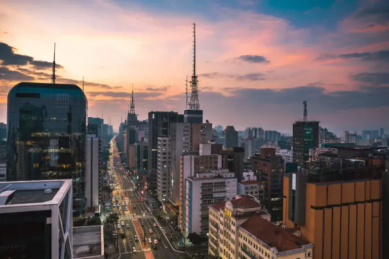 Imagem da paisagem urbana de São Paulo ao pôr do sol mostra uma avenida movimentada repleta de prédios altos e torres de rádio, com carros e postes de luz visíveis abaixo, sob um céu colorido com nuvens