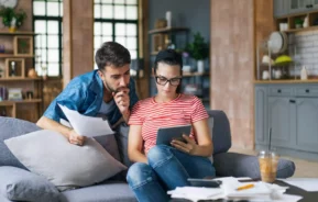 Imagem de uma mulher segurando um tablet sentada em um sofá que está na sala de um imóvel enquanto um homem está logo atrás apoiado na almofada do sofá com alguns papéis na mão para ilustrar matéria sobre taxa de financiamento imobiliário