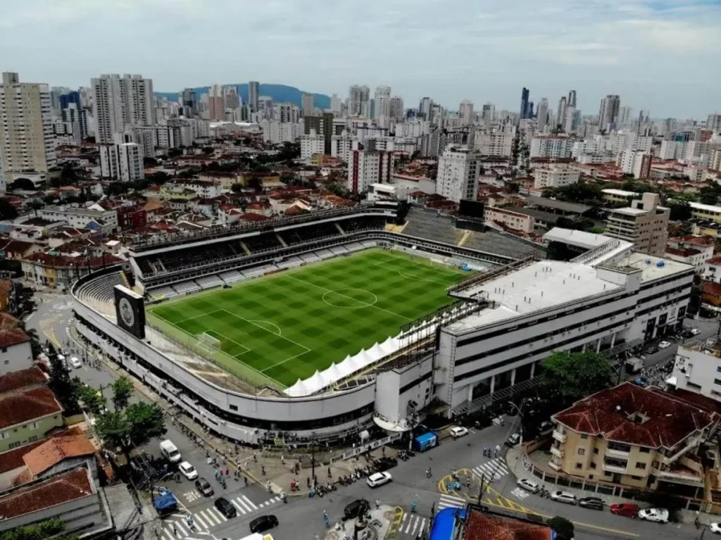 Imagem aérea de um grande estádio de futebol cercado por prédios e ruas na Vila Belmiro, em Santos, com grama verde no campo e áreas de assentos brancas em meio à paisagem urbana densamente povoada.