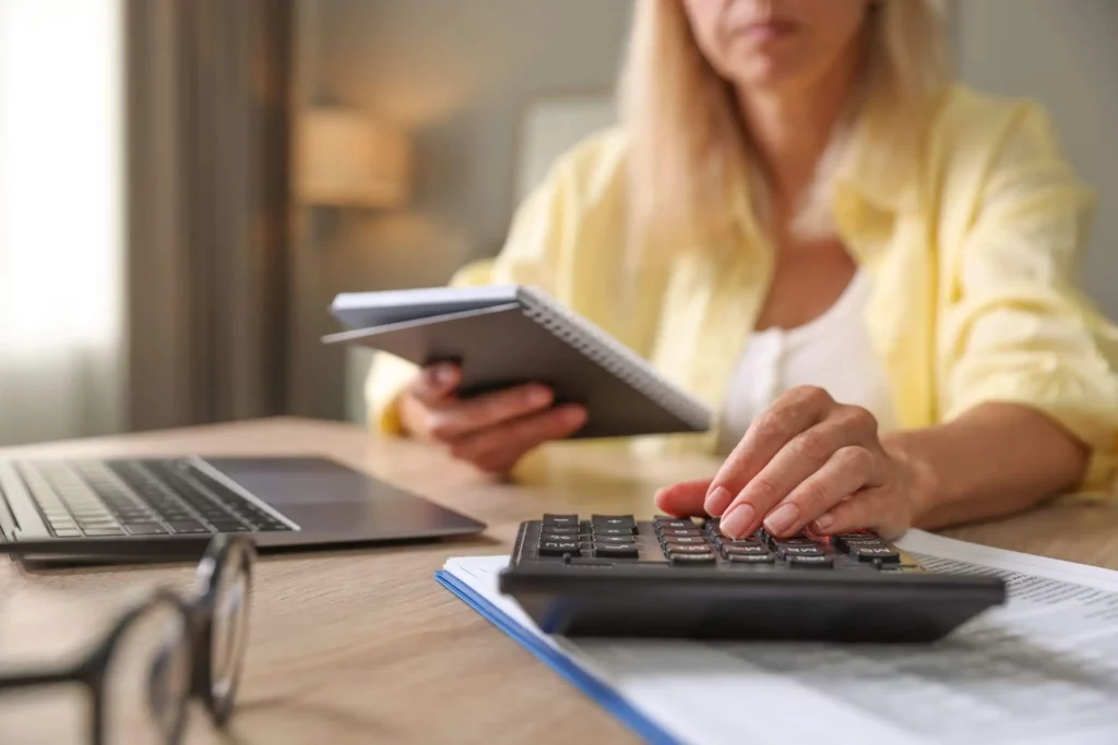 Imagem de uma mulher de camisa amarela sentada em uma mesa usando uma calculadora, com um caderno na mão. Um laptop, papéis e óculos sugerem que ela está concentrada em finanças ou orçamento