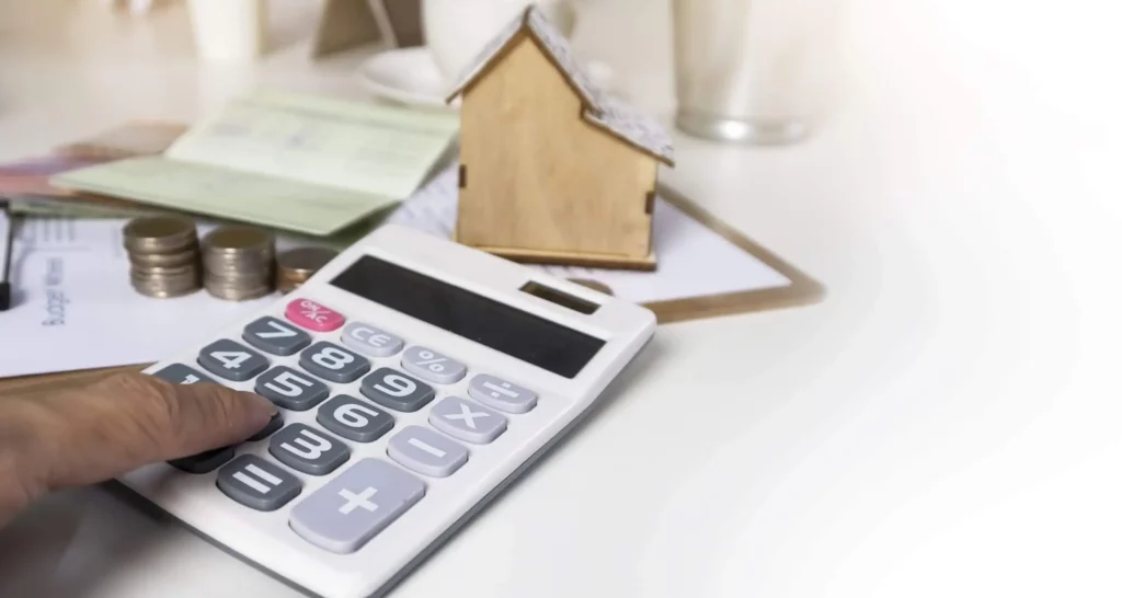 Imagem de uma mão pressionando botões em uma calculadora ao lado de pilhas de moedas, papéis e um pequeno modelo de casa de madeira