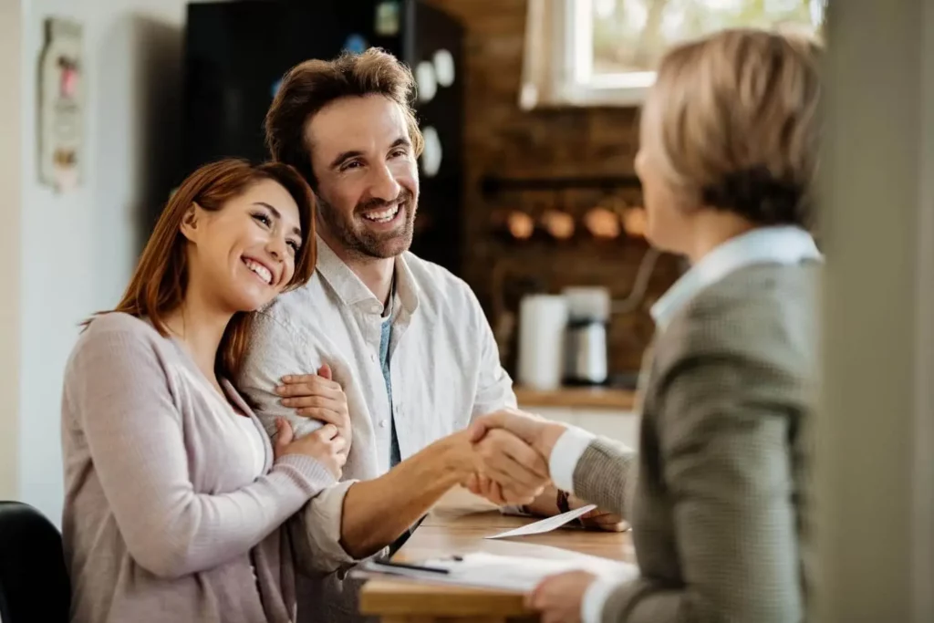 Imagem de um casal de um casal jovem sorrindo enquanto aperta as mãos de uma profissional para ilustrar matéria sobre como fazer um consórcio 