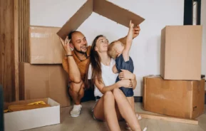 Imagem de uma família feliz de três pessoas está sentada no chão, entre caixas de papelão; os pais seguram a tampa de uma caixa sobre a cabeça, como se fosse um telhado, enquanto a criança aponta para cima, para ilustrar matéria que responde à pergunta: consórcio imobiliário vale a pena?