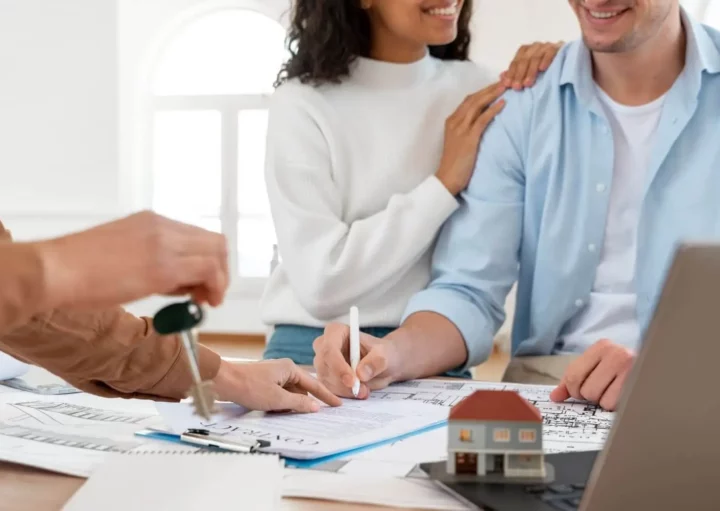 Imagem de um casal sorridente assinando um documento enquanto um agente imobiliário entrega uma chave para ilustrar matéria sobre consórcio imobiliário: vale a pena?